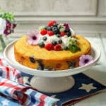 A round berry cake with visible blueberries, topped with whipped cream, fresh berries, and edible flowers, sits on a white cake stand over a patriotic-themed cloth.