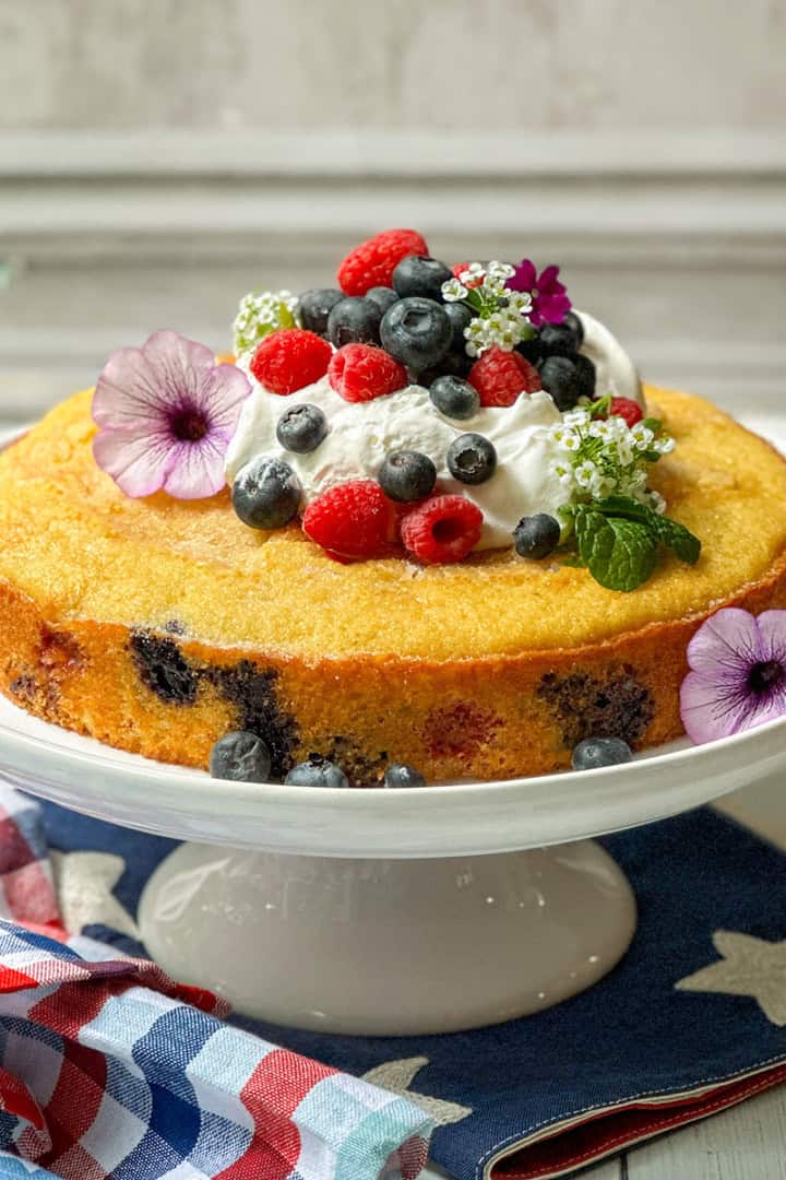 A round berry cake with visible blueberries, topped with whipped cream, fresh berries, and edible flowers, sits on a white cake stand over a patriotic-themed cloth.
