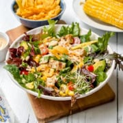 A bowl of southwest chicken salad with grilled chicken, avocado, cherry tomatoes, greens, yellow bell pepper, and sprouts on a wooden board, with corn on the cob and chips in the background.
