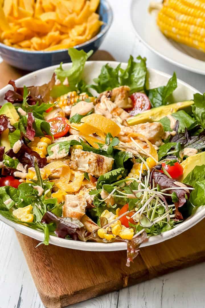 A bowl of southwest chicken salad with grilled chicken, avocado, cherry tomatoes, greens, yellow bell pepper, and sprouts on a wooden board, with corn on the cob and chips in the background.