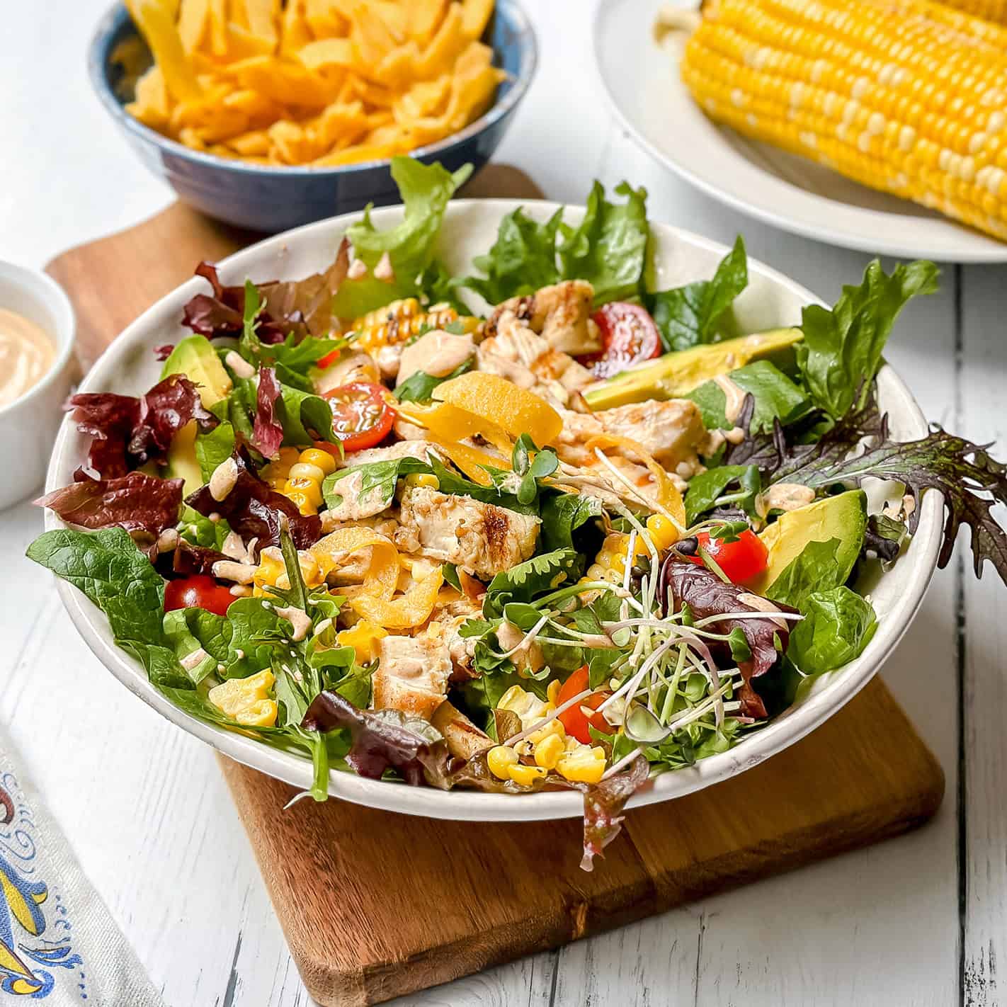 A bowl of southwest chicken salad with grilled chicken, avocado, cherry tomatoes, greens, yellow bell pepper, and sprouts on a wooden board, with corn on the cob and chips in the background.