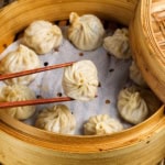 A bamboo steamer filled with steamed dumplings; one delicate morsel, made from a traditional Chinese dumplings recipe, is being held by chopsticks above the others.