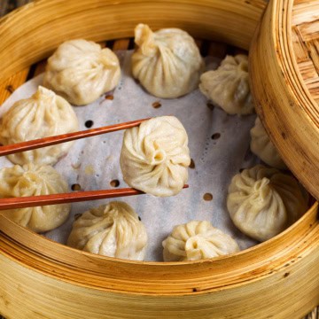 A bamboo steamer filled with steamed dumplings; one delicate morsel, made from a traditional Chinese dumplings recipe, is being held by chopsticks above the others.