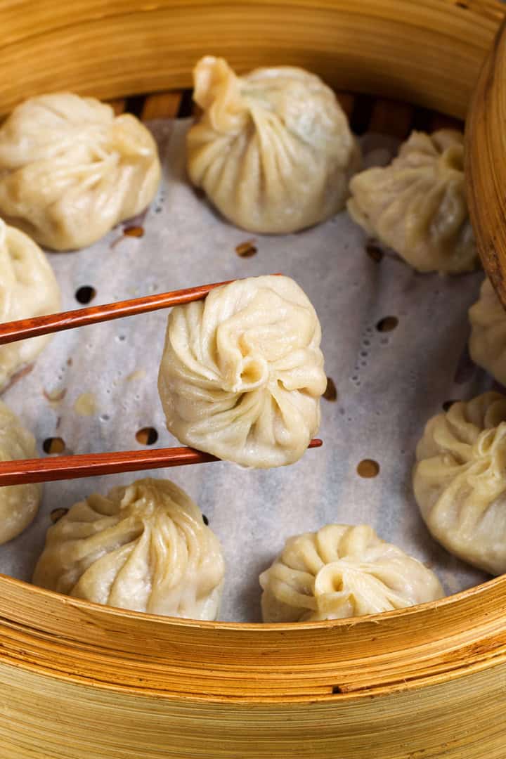 A bamboo steamer filled with steamed dumplings; one delicate morsel, made from a traditional Chinese dumplings recipe, is being held by chopsticks above the others.