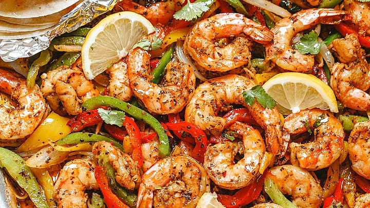 Sheet pan of cooked shrimp with sliced red, yellow, and green bell peppers, garnished with lemon wedges and cilantro, served alongside a stack of tortillas wrapped in foil—perfect for easy May 30 Minute Dinners.