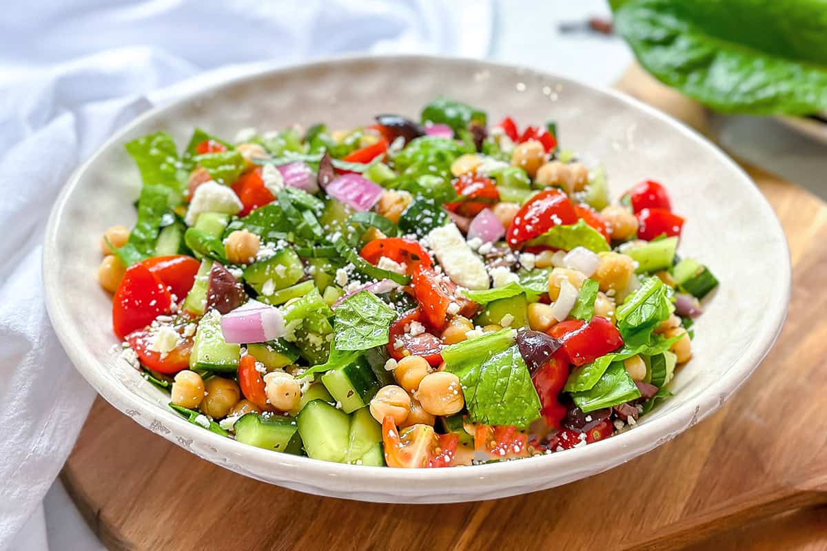A bowl of Mediterranean Chopped Salad with lettuce, cucumbers, tomatoes, red onions, chickpeas, olives, and crumbled cheese sits on a wooden surface.