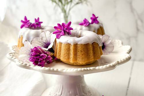 Horizontal side view of Lavender Earl Grey Tea Cake on a white serving stand with fresh lilacs and pansies.