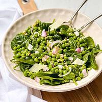 Horizontal view of pea salad in a white bowl with serving utensils on a round wood board.