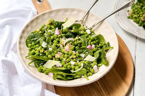 Horizontal view of pea salad in a white bowl with serving utensils on a round wood board.