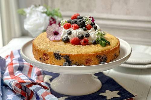 Side view of unsliced berry cake on a white cake stand and topped with Chantilly Cream.