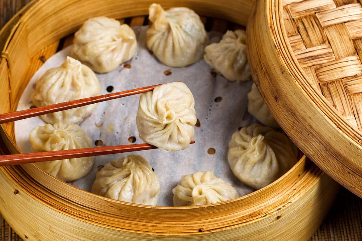 A pair of chopsticks holds a delicate steamed dumpling above a bamboo steamer filled with similar treats on parchment paper, capturing the irresistible appeal of an authentic chinese dumplings recipe.