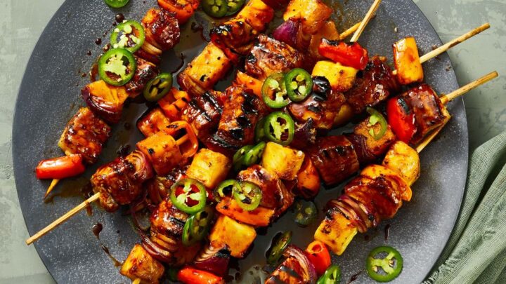 Five skewers with grilled vegetables, pineapple, and meat are arranged on a dark plate, garnished with sliced jalapeños—perfect for Healthy High Protein Dinners—next to a small bowl of sauce and a green napkin.