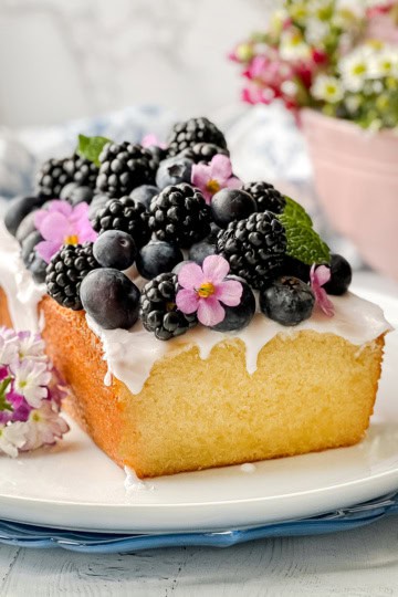 A rectangular pound cake with white icing, topped with blackberries, blueberries, mint leaves, and pink flowers, sits on a white plate next to a small cluster of blossoms.