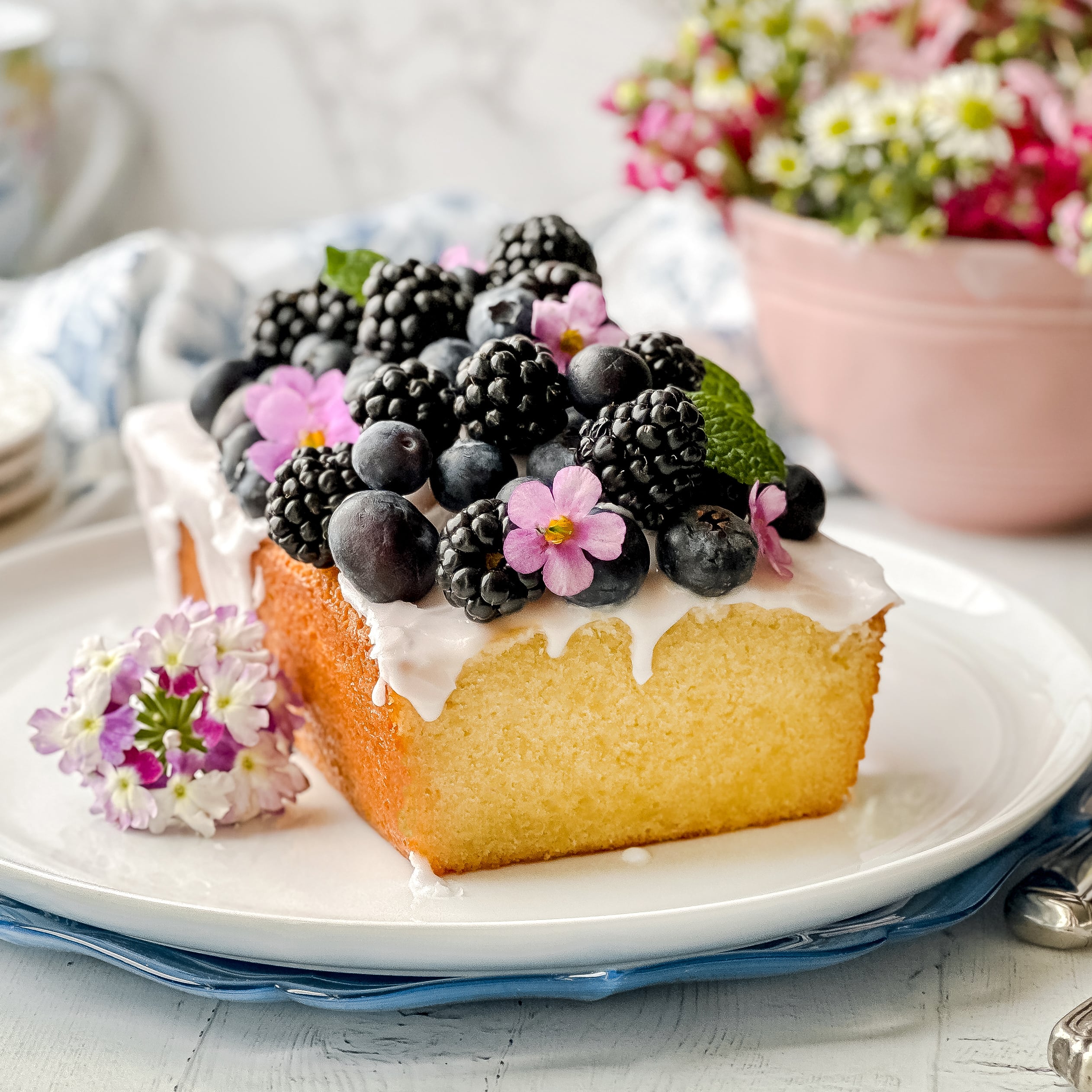 A loaf pound cake with white icing, topped with blackberries, blueberries, small pink flowers, and mint leaves, is served on a white plate with a purple flower garnish.