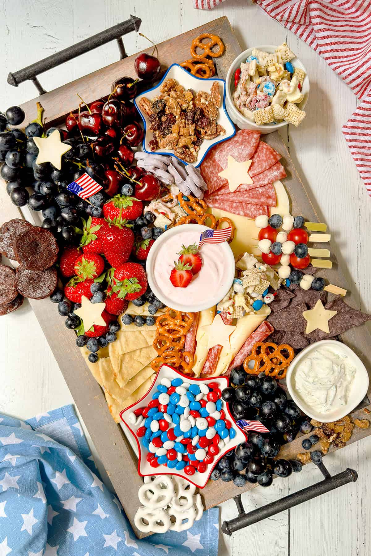 A festive Fourth of July Snack Board featuring assorted snacks—fruit, cheese, meats, crackers, pretzels, yogurt, snack mix, and candies in red, white, and blue—decorated with small American flags for a patriotic touch.