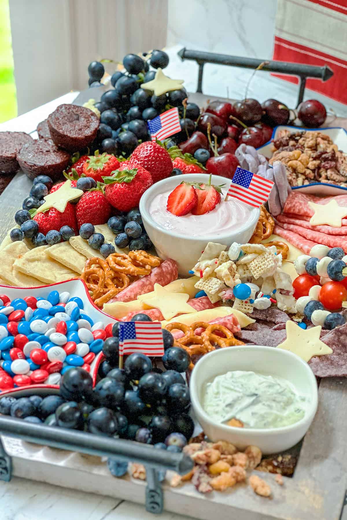 A patriotic-themed Fourth of July Snack Board featuring red, white, and blue treats, fruits, pretzels, dips, cheese, and festive American flag toothpicks.