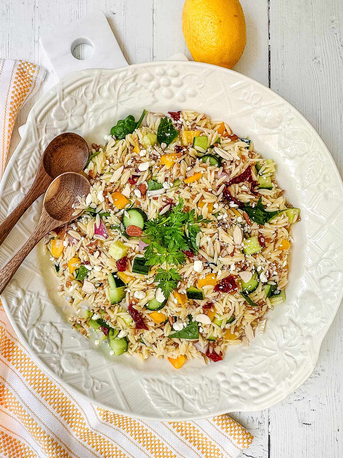 A large white plate of orzo and farro salad with chopped vegetables, herbs, and feta cheese, accompanied by two wooden spoons and a lemon on a white wooden surface.
