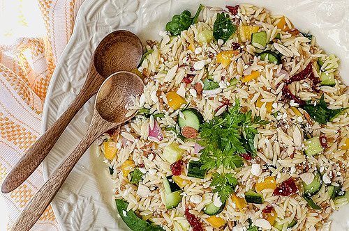A plate of orzo pasta salad with vegetables, feta cheese, and herbs, served with three wooden spoons on a white table next to a yellow-striped cloth-perfect for fans of farro salad looking for a fresh twist.