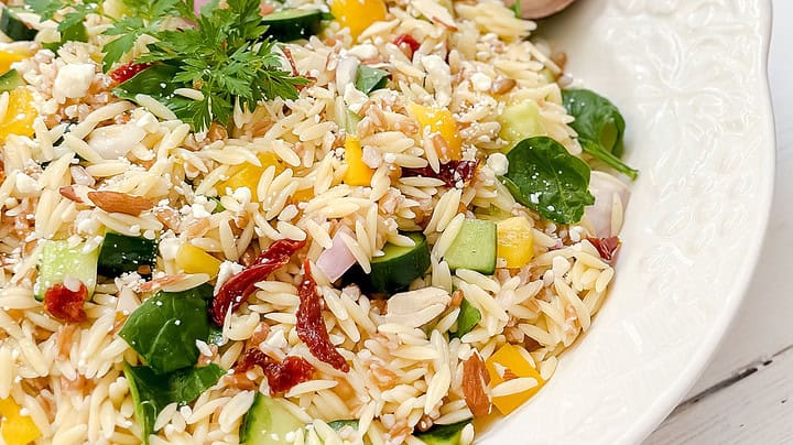A white bowl filled with orzo salad—similar to a farro salad—containing spinach, cucumber, yellow bell pepper, red onion, sun-dried tomatoes, feta cheese, and parsley, with two wooden serving spoons.