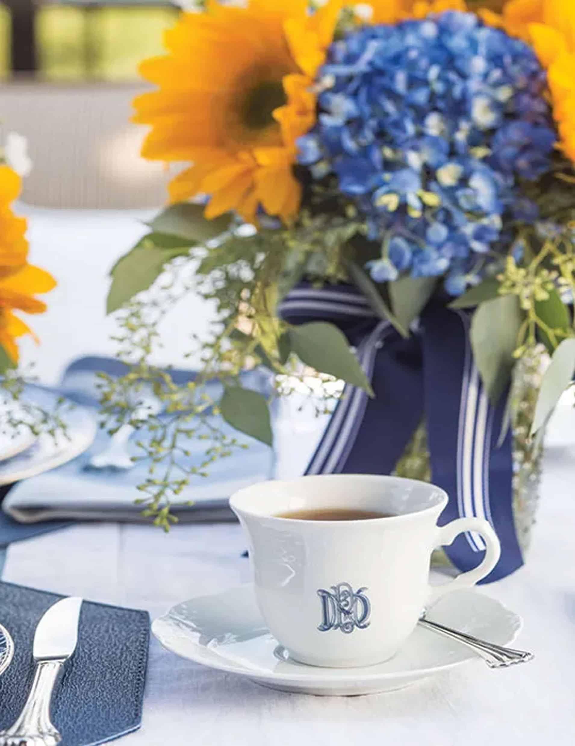 A white teacup with a monogram sits on a saucer beside a spoon, with a bouquet of sunflowers and blue hydrangeas in the background-a charming setting for a Father's Day tea party.