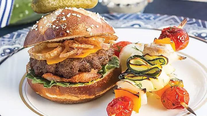 Side view of mini cheeseburgers with fried shallots on a bun with a skewer of roasted vegetables for an outdoor Father's Day afternoon tea.