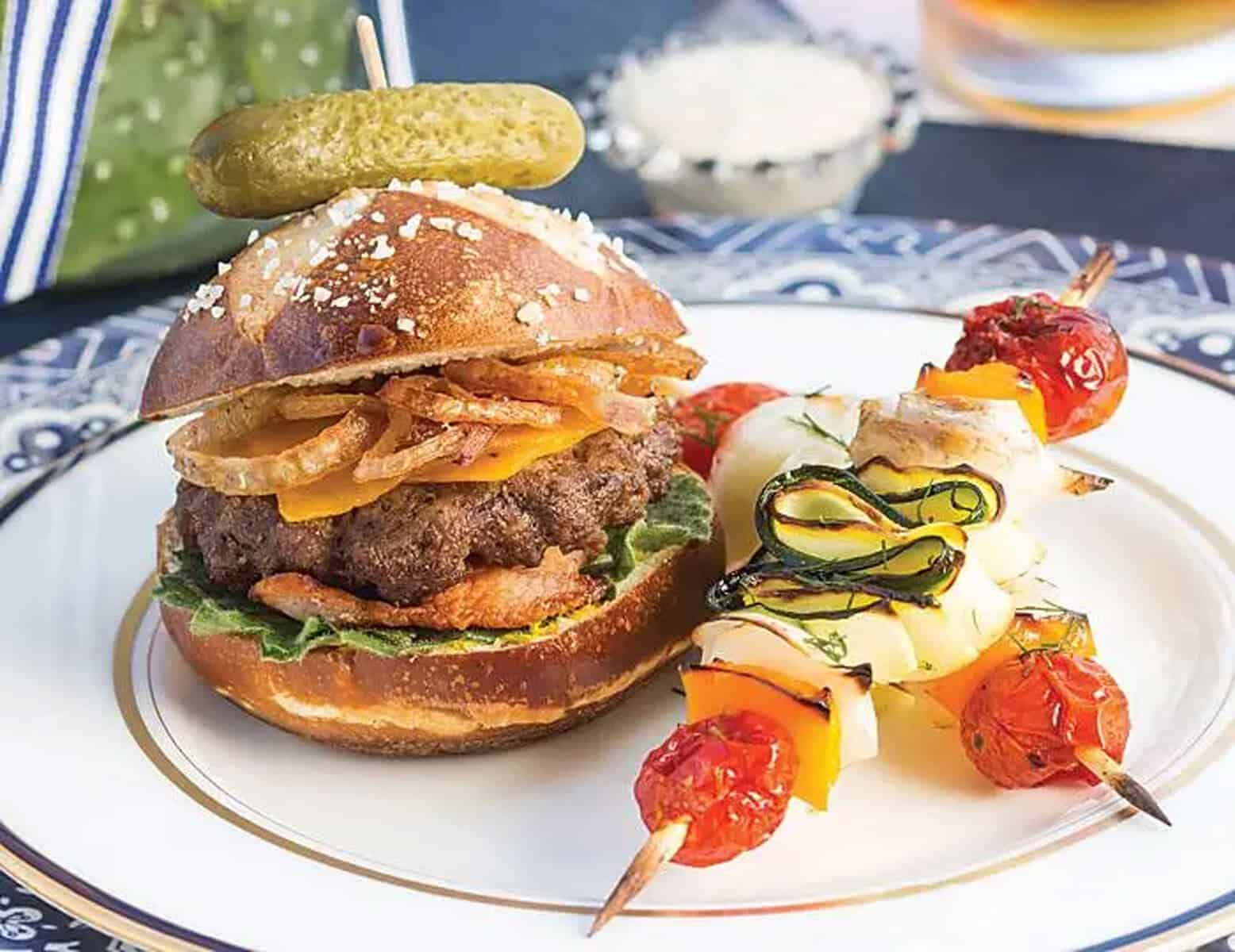 Side view of mini cheeseburgers with fried shallots on a bun with a skewer of roasted vegetables for an outdoor Father's Day afternoon tea.