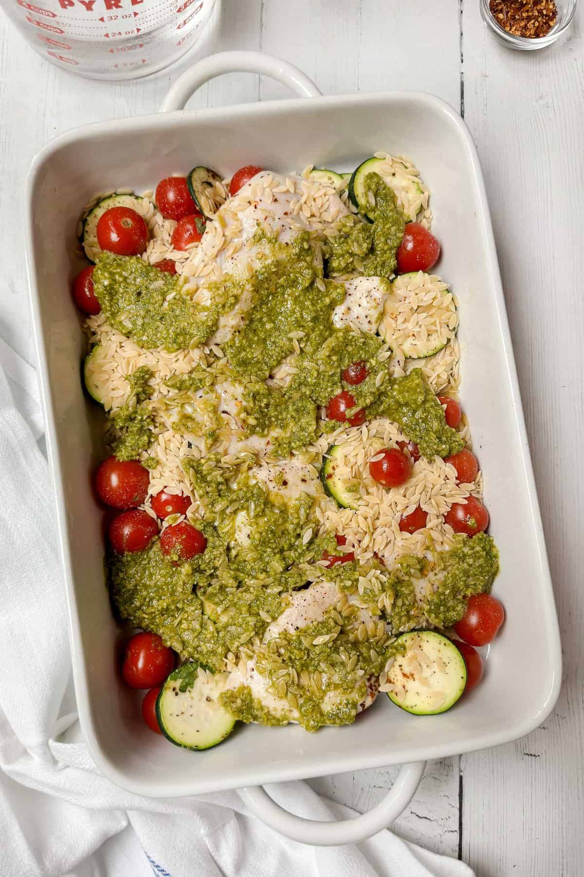 Top view of dry orzo and pesto added to white casserole dish.