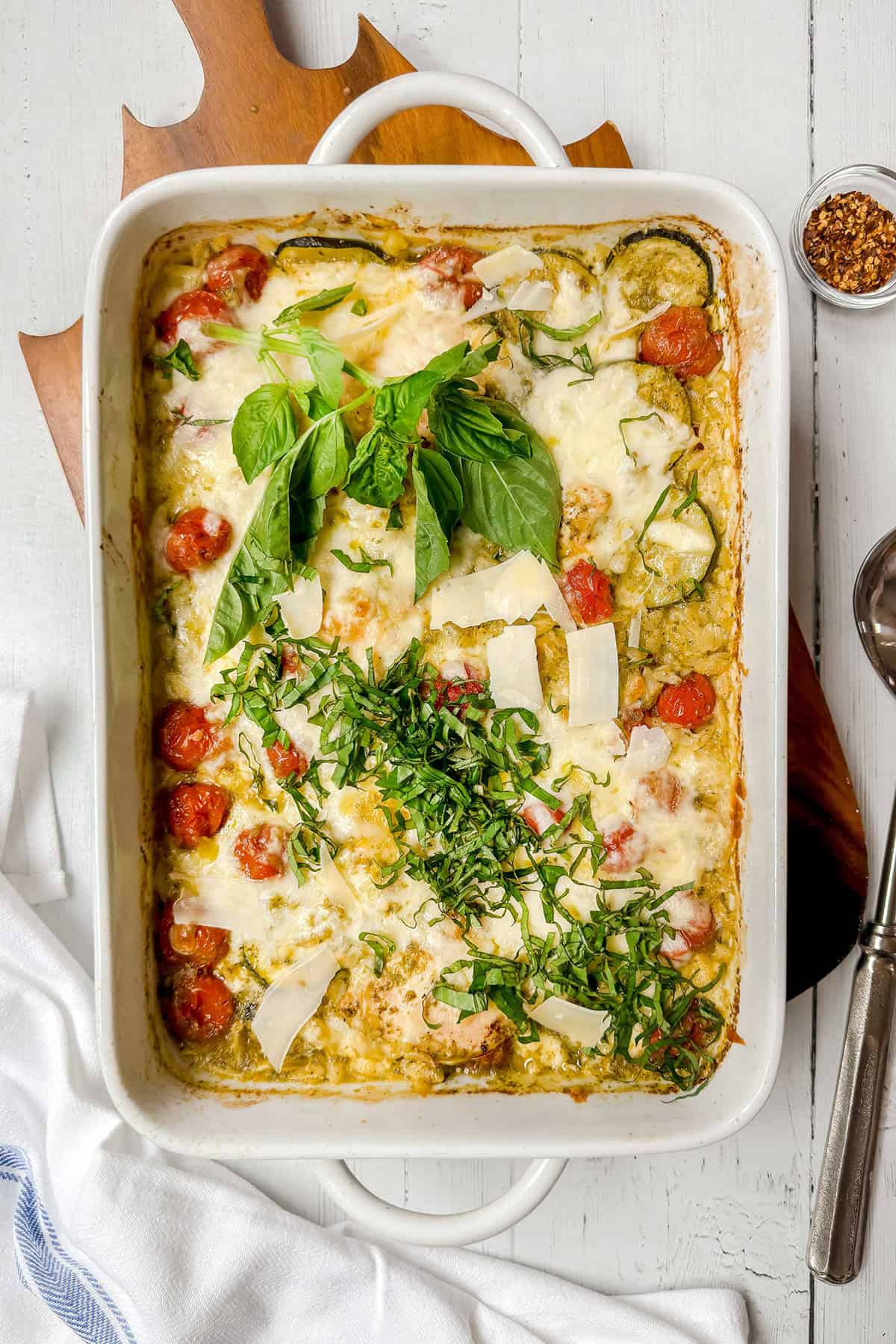 Adding cheese and fresh herbs to Pesto Caprese Chicken in a rectangular baking dish.