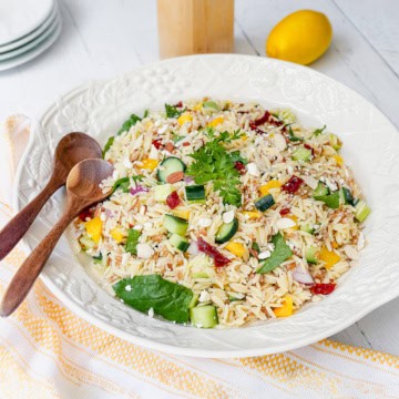 A bowl of orzo salad with cucumber, spinach, yellow peppers, sun-dried tomatoes, parsley, and feta cheese sits on a white table alongside wooden spoons, a lemon, and plates-perfect for fans of orzo or farro salad.