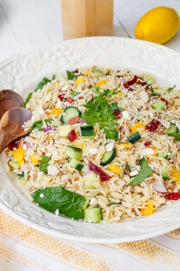 A bowl of orzo salad with cucumber, spinach, yellow peppers, sun-dried tomatoes, parsley, and feta cheese sits on a white table alongside wooden spoons, a lemon, and plates-perfect for fans of orzo or farro salad.