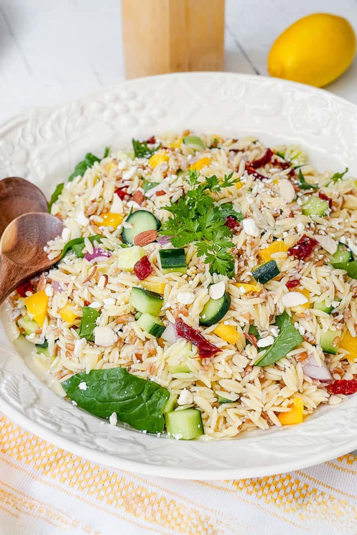 A bowl of orzo salad with cucumber, spinach, yellow peppers, sun-dried tomatoes, parsley, and feta cheese sits on a white table alongside wooden spoons, a lemon, and plates-perfect for fans of orzo or farro salad.