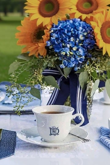 A white table set outdoors for a Father's Day tea party, featuring cups of coffee, plates, silverware, and a centerpiece of sunflowers, blue hydrangeas, and greenery tied with a blue ribbon.