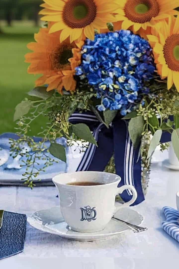A white table set outdoors for a Father's Day tea party, featuring cups of coffee, plates, silverware, and a centerpiece of sunflowers, blue hydrangeas, and greenery tied with a blue ribbon.