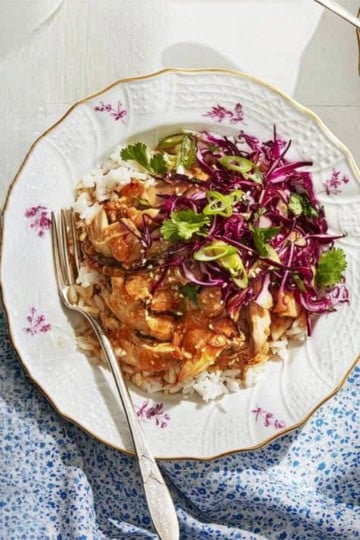A plate of rice topped with saucy chicken, red cabbage slaw, and cilantro-an effortless summer slow cooker meal-sits on a white table with a blue-patterned cloth and a fork resting on the plate.