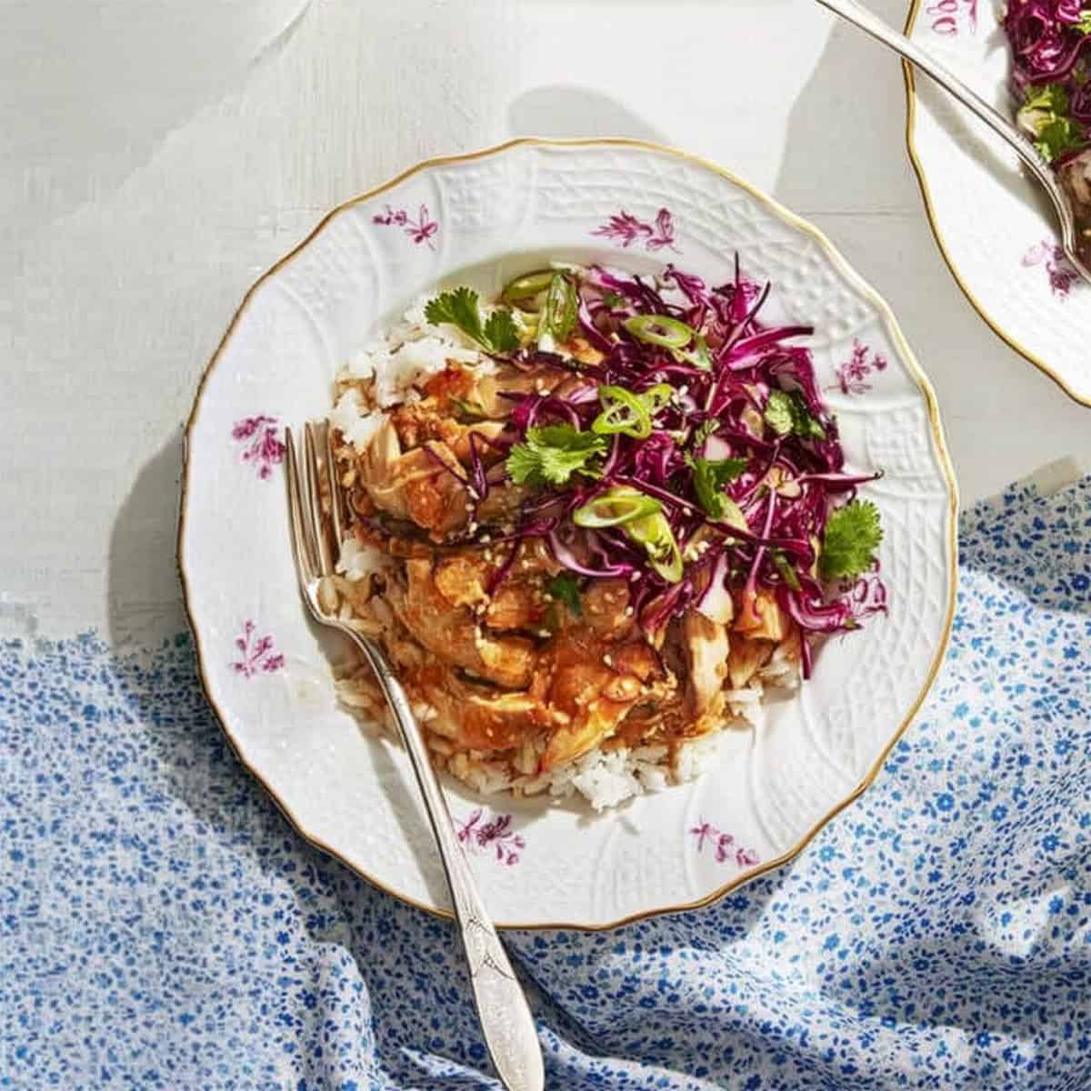 A plate of rice topped with saucy chicken, red cabbage slaw, and cilantro—an effortless summer slow cooker meal—sits on a white table with a blue-patterned cloth and a fork resting on the plate.