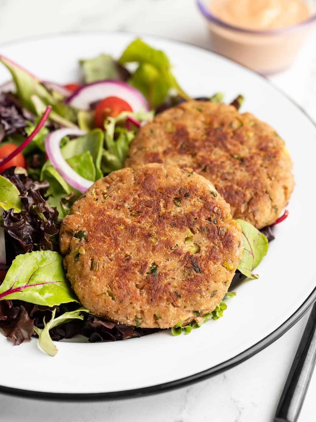 Two golden-brown salmon patties sit on a bed of mixed greens with sliced red onion and cherry tomatoes, served on a white plate-a perfect choice for healthy, budget friendly dinners.