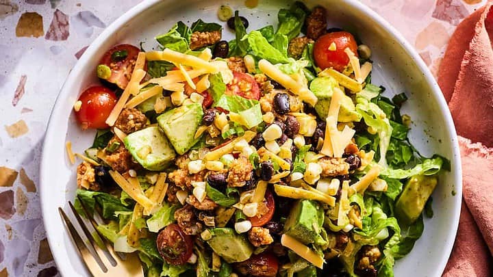 A bowl of salad with lettuce, cherry tomatoes, avocado, black beans, shredded cheese, and a fork—perfect for June 30 Minute Dinners—sits next to a small bowl of green dressing and a glass of pink beverage.