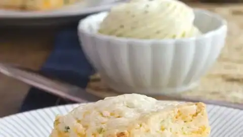 A cheddar and chive biscuit sits on a white plate, perfect for a Father's Day tea party, with a bowl of whipped butter and another biscuit in the background.