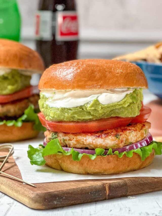Side view of Chicken Burger topped with guacamole, tomatoes, lettuce, and red onion on a wood board for serving.