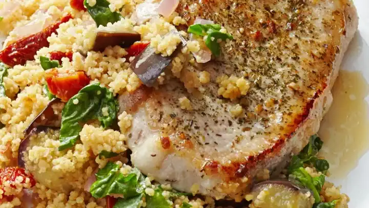 A seasoned pork chop served on a bed of couscous with spinach, eggplant, and sun-dried tomatoes—perfect for quick June 20 Minute Dinners—presented beautifully on a white plate.