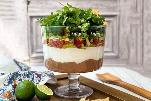 Horizontal view of easy 7 Layer Dip in a glass trifle dish with tortilla chips.