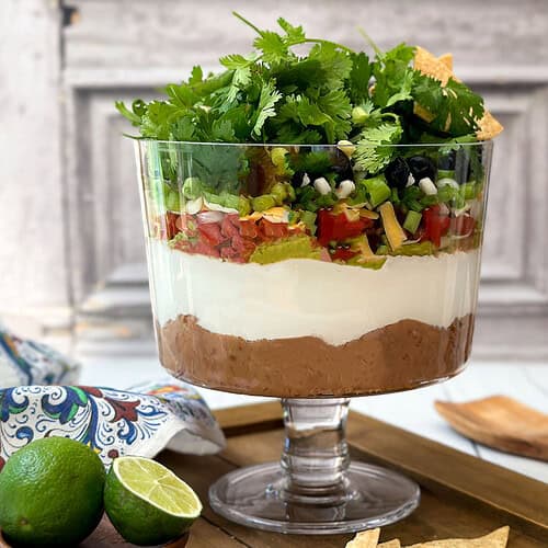 Horizontal view of easy 7 Layer Dip in a glass trifle dish with tortilla chips.