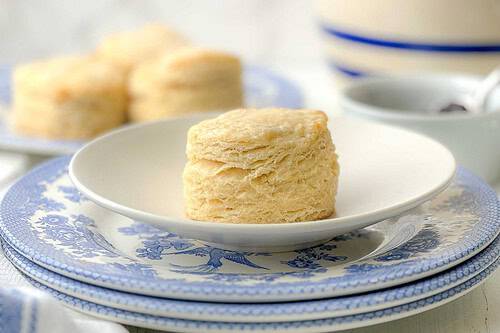 A flaky biscuit from a classic biscuit recipe sits on a white plate with blue floral trim, with more biscuits and a cup blurred in the background.