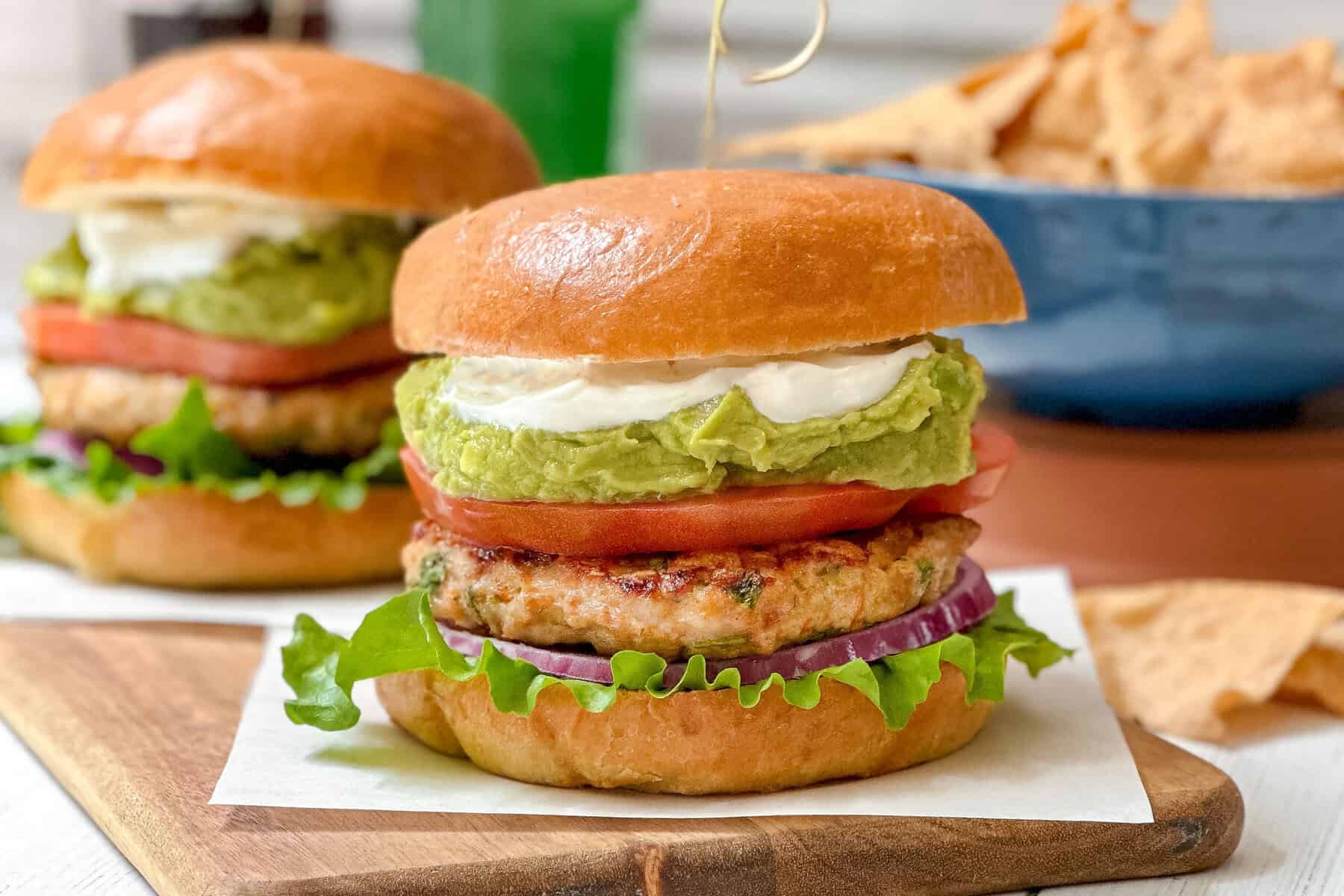 Horizontal side view of Cheddar Chicken Burger in a bun with guacamole, sour cream, tomato slices and onion slices.