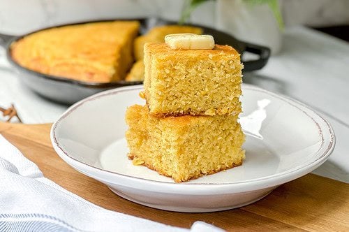 Side horizontal view of two stacked squares of cornbread with butter on a white plate.