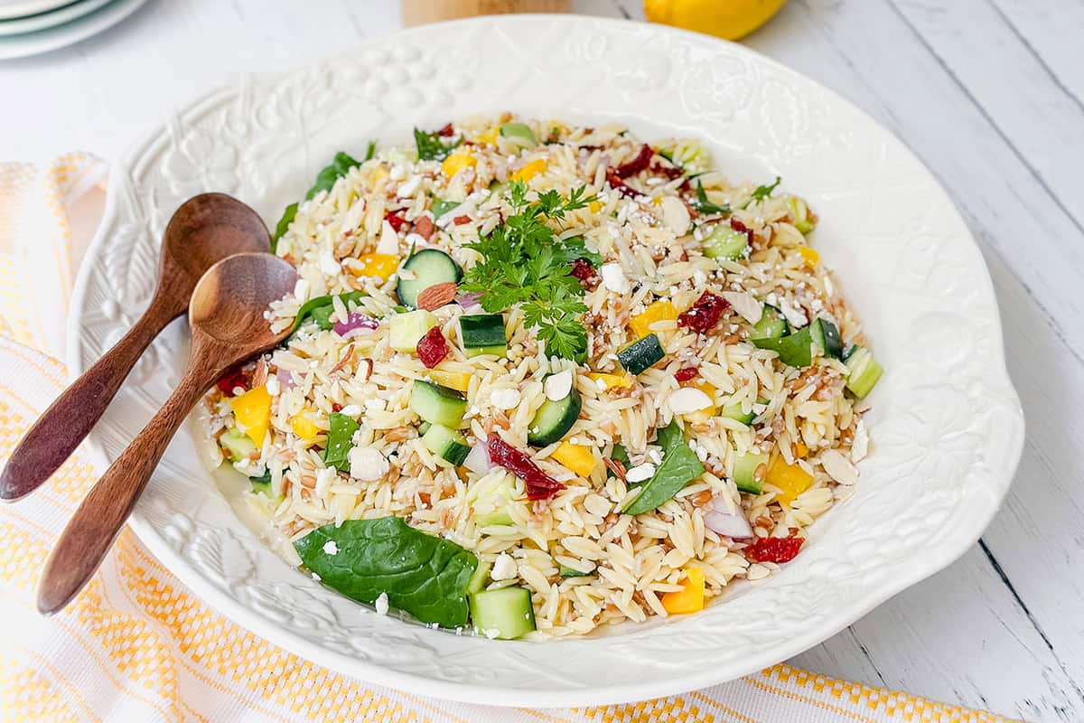 A large white bowl of farro pasta salad with cucumber, bell pepper, spinach, sun-dried tomatoes, cheese, and parsley is served alongside two wooden spoons-perfect for fans of vibrant farro salad.