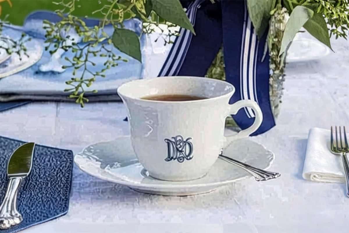 A white teacup with a monogram sits on a matching saucer with a spoon, placed on a table set for a Father's Day tea party, complete with silverware and decorated with a floral arrangement.