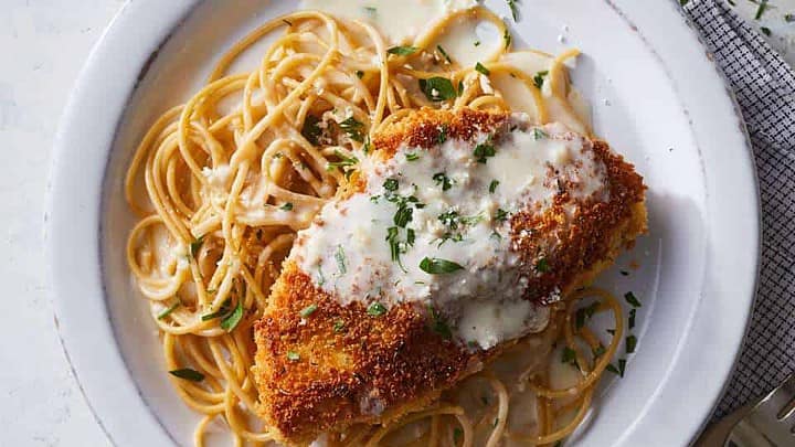 Top view of chicken parm over whole grain spaghetti with fresh herbs on a white round plate.