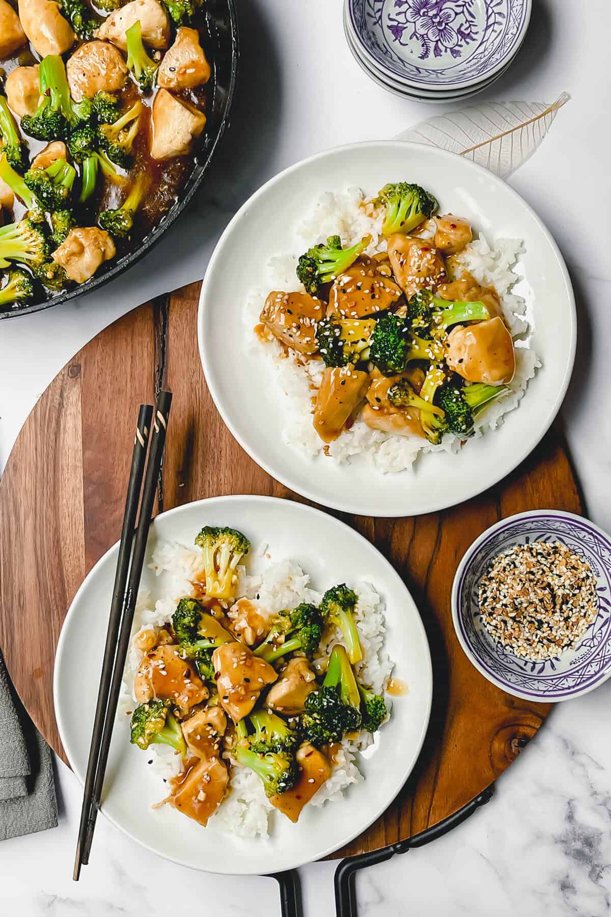 Two plates of Chicken and Broccoli over white rice are topped with a savory sauce and sesame seeds. Chopsticks rest on one plate, while a skillet and a bowl of seasoning sit nearby on a wooden board.