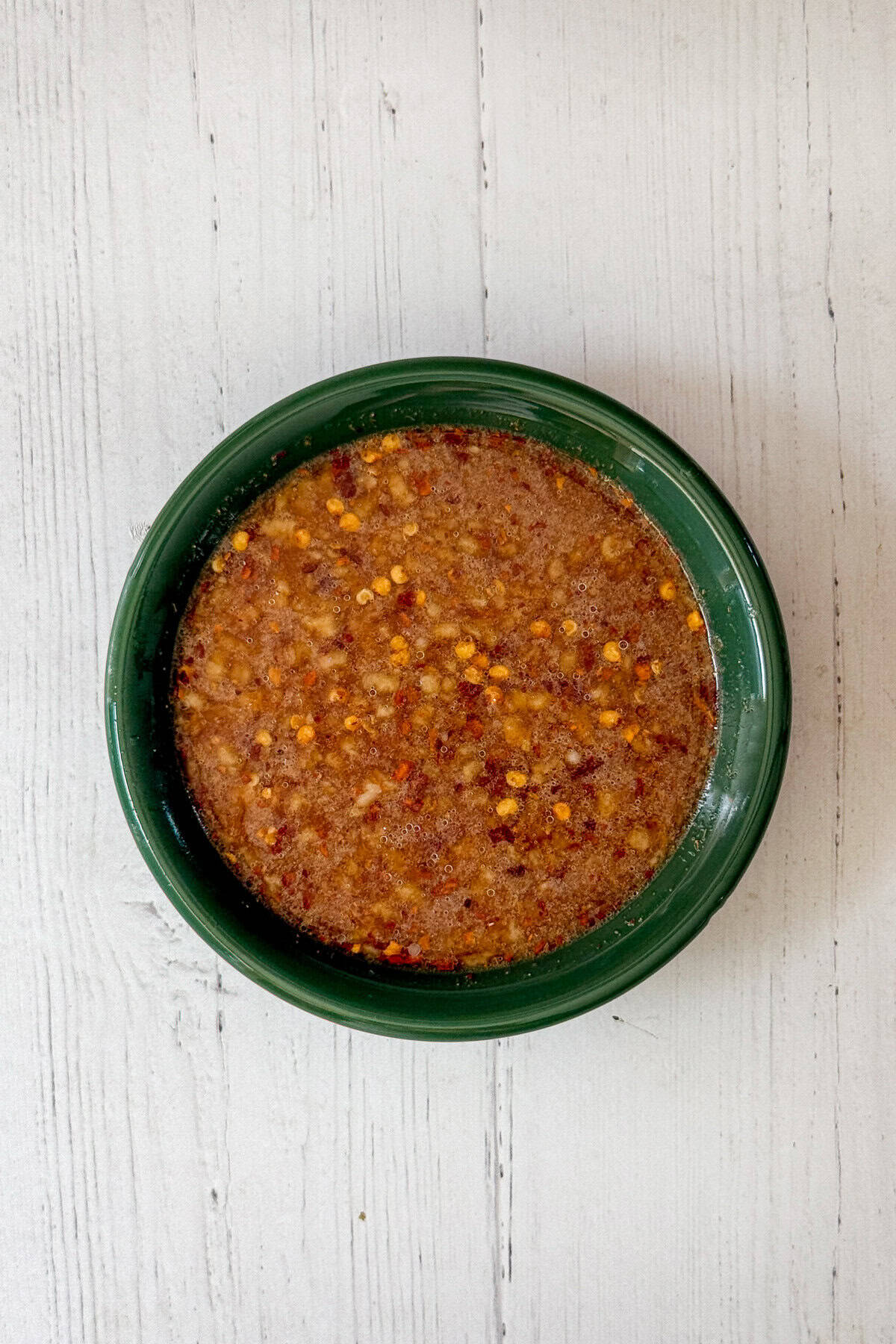 A green bowl filled with a brown, oily chicken and broccoli sauce speckled with red pepper flakes, placed on a white wooden surface.